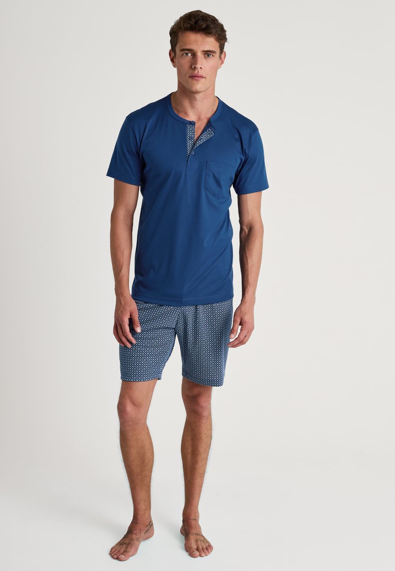 Korte mouwen, marineblauwe shirt met een gebloemd knoopjespatroon en zak, gecombineerd met gebloemde blauwe shorts. Beide gemaakt van lichtgewicht stof.