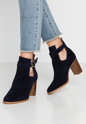 Persoon draagt zwarte suède enkel laarzen met houten blokhakken en gouden gespbanden, gecombineerd met lichtblauwe jeans met rafelige zoom.