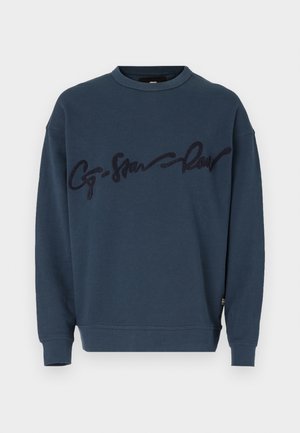 Mørkeblå genser med rund hals og ribbede mansjetter, med en opphøyet mørkeblå skriftlogo over brystet. Myk tekstur.