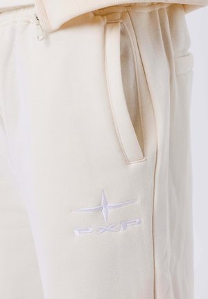 Pantaloni sportivi beige con una texture morbida, dotati di tasche laterali e un logo ricamato bianco "PXP" vicino alla tasca sinistra.