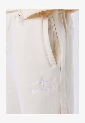 Pantaloni sportivi beige con una texture morbida, dotati di tasche laterali e un logo ricamato bianco "PXP" vicino alla tasca sinistra.