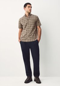 Man standing with hands in pockets wearing a beige plaid short-sleeve button-up shirt, dark pants, and dark brown shoes on white background.