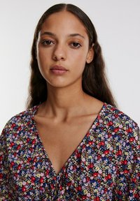 Blumengemustertes Kleid mit tiefem V-Ausschnitt, kurzen Ärmeln und einem mehrfarbigen Design in Rot-, Violett- und Grüntönen auf dunklem Hintergrund.