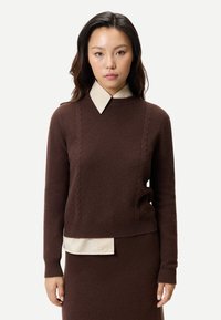 Ung kvinna med långt mörkt hår bär en brun stickad tröja över en beige skjorta med krage och en matchande brun kjol, stående mot en vit bakgrund.