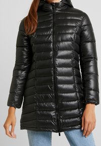 Žena v černé prošívané dlouhé puffrové bundě s předním zipem a kapucí, v kombinaci se světle modrými džíny.