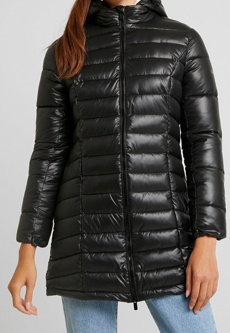 Žena v černé prošívané dlouhé puffrové bundě s předním zipem a kapucí, v kombinaci se světle modrými džíny.