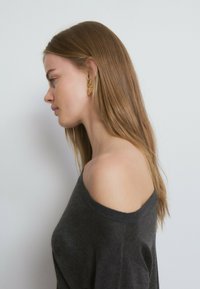 Suéter gris carbón con hombros descubiertos, textura suave y ajuste relajado. Pendientes de hoja dorada que aportan detalle, con cabello largo y liso cayendo hacia abajo.