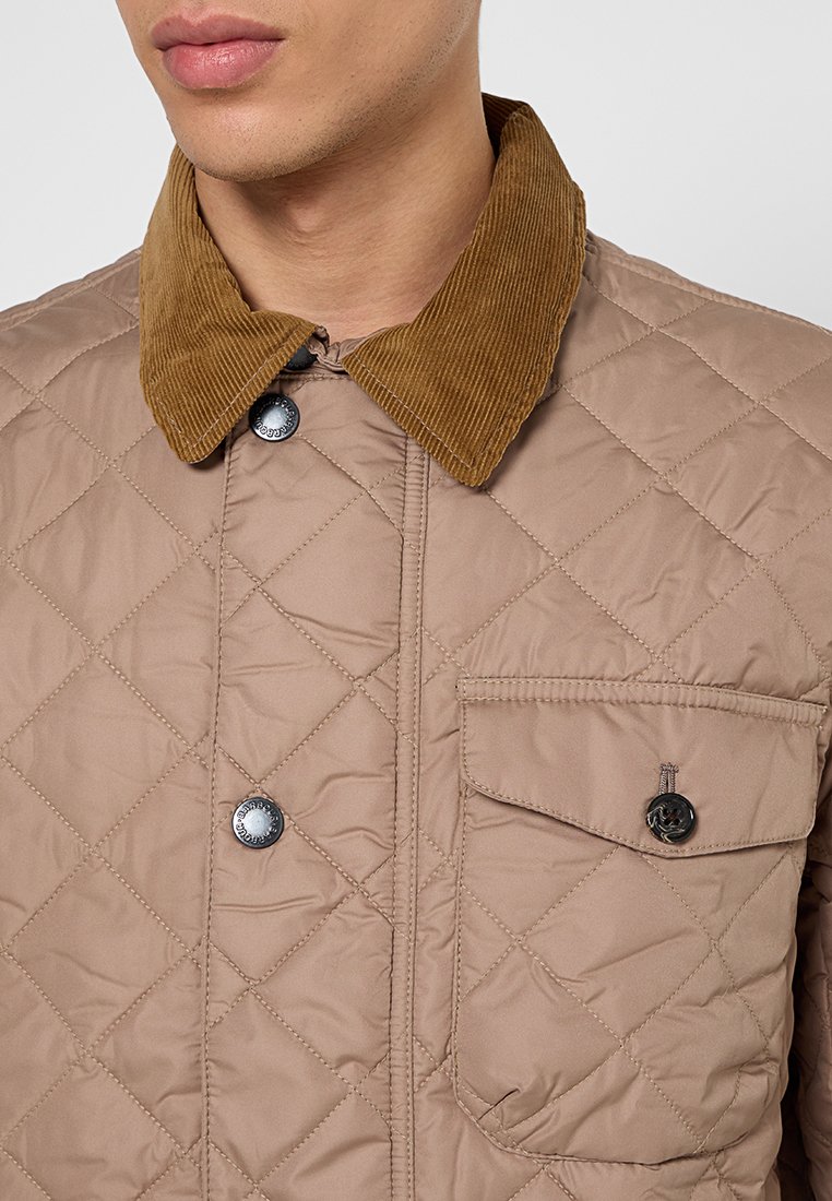 Veste beige matelassée avec un col en velours côtelé marron, ornée de boutons noirs et d'une poche poitrine. Texture lisse avec un motif en losange.