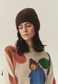 Rjava pletena kapa z rebrastim vzorcem. Bež pulover z barvitimi cvetličnimi in medvedjimi motivi. Teksturirana tkanina z opaznimi akcenti.