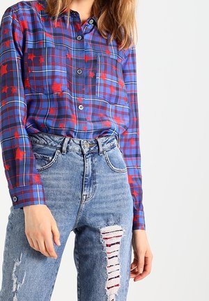 Kvinde iført en blå ternet skjorte med røde stjerner stoppet ned i højttaljede, revnede blå jeans mod en hvid baggrund.