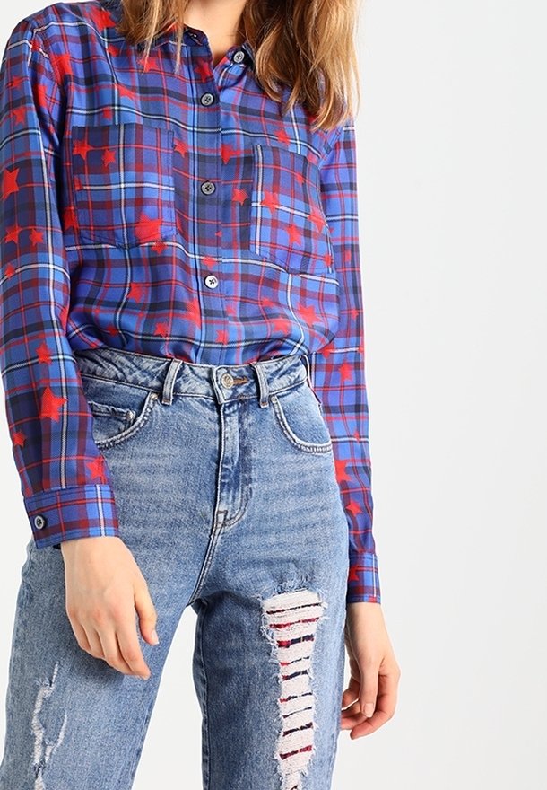 Femme portant une chemise à carreaux bleue avec des étoiles rouges rentrée dans un jean bleu déchiré taille haute, sur fond blanc.