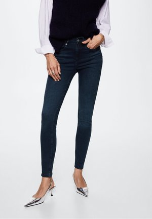 Vrouw draagt donkerblauwe skinny jeans, witte blouse met opgerolde mouwen, zwarte mouwloze trui en zilveren slingback schoenen met hoge hak.