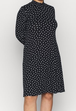 Black knee-length dress with white polka dots, long sleeves, and a high ruched neckline, worn by a person with hands behind their back.