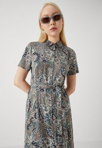 Button-up dress with a paisley pattern in shades of teal, navy, and cream, featuring short sleeves and a tie belt at the waist.