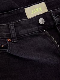 Jeans de mezclilla negra con cremallera y cierre de botón visibles. Entre sus características se incluye una etiqueta de tela con "JJXX" en amarillo brillante y remaches de cobre.