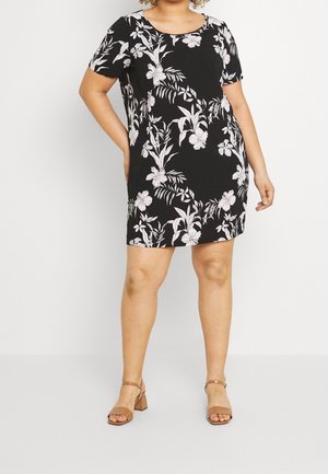 Kobieta ubrana w czarną sukienkę z krótkim rękawem z białym kwiatowym wzorem oraz beżowe sandały na słupku z odkrytymi palcami, stojąca z jedną ręką na biodrze.