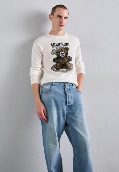 Vit stickad tröja med björngrafik och "MOSCHINO"-text, tillsammans med ljusblå baggy jeans, som visar en avslappnad passform och avslappnad stil.