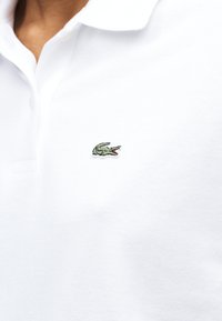 Άσπρο πόλο μπλουζάκι με πράσινο λογότυπο κροκόδειλου κεντημένο στο στήθος, με ανοιχτό γιακά και υφασμάτινη υφή.