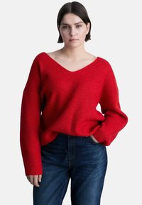 Maglione rosso a maglia con scollo a V e maniche lunghe. Presenta una vestibilità comoda e una texture a coste. Abbinato a jeans in denim scuro.