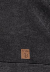 Tejido de punto gris oscuro con una apariencia texturizada, que presenta una etiqueta cuadrada de cuero marrón con la inscripción "KEY LARGO" y un diseño de logo en relieve.