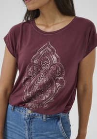 T-shirt bordeaux à manches courtes en tissu doux avec un motif paisley blanc détaillé sur le devant, associé à un jean en denim bleu clair.