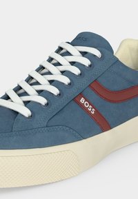 Baskets en daim bleu avec lacets blancs, semelle crème et bande latérale rouge comportant le mot « BOSS » en lettres blanches.