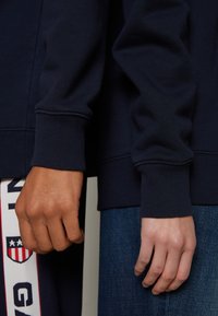 Due mani che indossano felpe di colore blu scuro. Una manica presenta polsini a coste, mentre l'altra è liscia con una cucitura visibile.