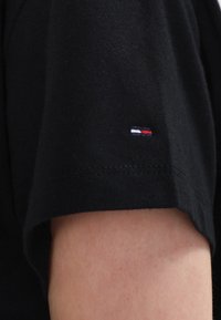 Černé tričko s krátkým rukávem na paži, s malým obdélníkovým logem s horizontálními bílými, modrými a červenými pruhy blízko lemu.