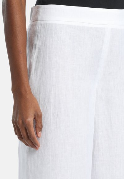 Gros plan sur une personne portant un pantalon en lin blanc et un haut noir, montrant la taille, la hanche et la main gauche détendue le long du corps.