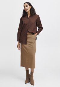 Bruine button-up shirt over een aansluitende beige kokerrok, gecombineerd met bijpassende beige enkellaarsjes. Eenvoudige, gladde texturen door het geheel.