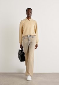 Polo a maniche lunghe beige con bottoni, abbinato a jeans beige chiaro. Il modello porta una borsa nera e indossa sneakers bianche.