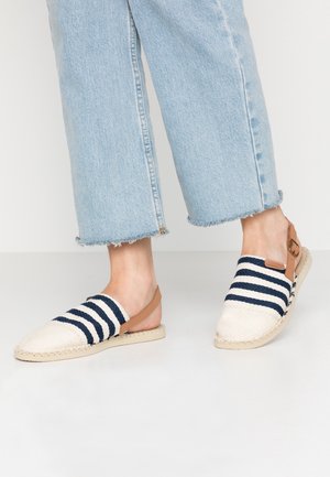 Fødder iført blå- og hvidstribede slip-on espadrille-sandaler med brune ankelremme og lyseblå korte jeans med frynsete kanter.