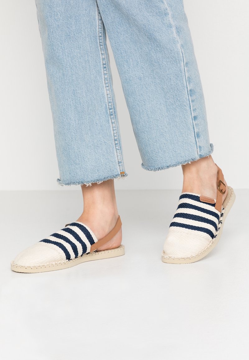Pieds portant des sandales espadrilles slip-on rayées bleu et blanc avec des brides de cheville couleur sable et un jean court bleu clair aux ourlets effilochés.