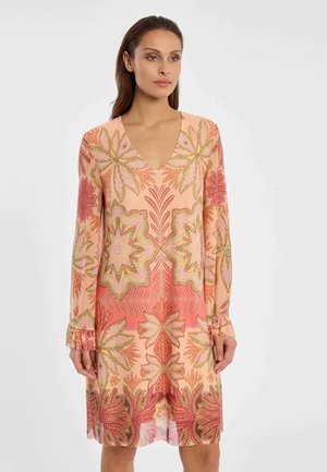 Femme portant une robe à manches longues et col en V avec des motifs floraux roses, orange et verts sur un fond blanc uni.