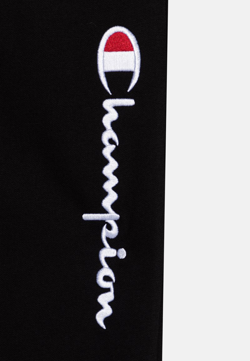 Gamba in stoffa nera con ricamo bianco "Champion"; il logo presenta un accento rosso, bianco e nero in una forma circolare sopra il testo.