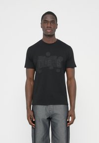 Μαύρο μπλουζάκι με υφασμένο λογότυπο «Guess» σε σκούρους τόνους. Κοντά μανίκια, στρογγυλή λαιμόκοψη, κατασκευασμένο από μαλακό, ελαφρύ ύφασμα.