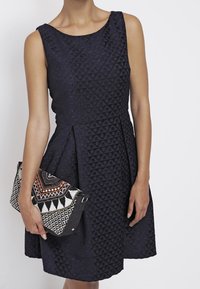 Granatowa sukienka o fakturowanej strukturze z dopasowanym krojem i okrągłym dekoltem, w zestawieniu z wzorzystą torebką kopertową w czerni i bieli z pomarańczowymi akcentami.