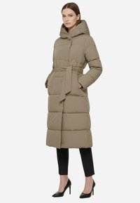 Parka khaki imbottita con cappuccio, vita a cintura, texture trapuntata, lunghezza lunga e tasche laterali, abbinata a pantaloni neri slim e tacchi.