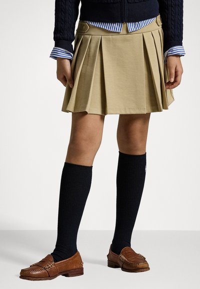 Pleedirätiku khaki seelik külgniitidega, millele on lisatud tumesinine nokakudumine särk ja siniste triipudega särk, mustad kuni põlveni sokid ja pruuni värvi loaferid.