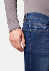 Dunkelblaue Jeans aus Denim mit ausgewaschenen Akzenten und aufgesteppten Taschen. Eine Hand ruht auf dem Hosenbund, der ein gewebtes Stoffarmband trägt.