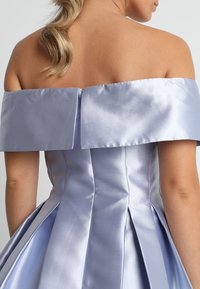 Žena s blond vlasy v culíku, oblečená v světle modrých saténových šatech s odhalenými rameny a plisovanou sukní, zobrazená zezadu.