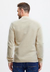 Beige gyapjú pulóver kerek nyakkivágással, bordázott mandzsettákkal és szegéllyel. A textúra kézzel fogható; a hátsó nézet a design és a szín egyszerűségét mutatja.