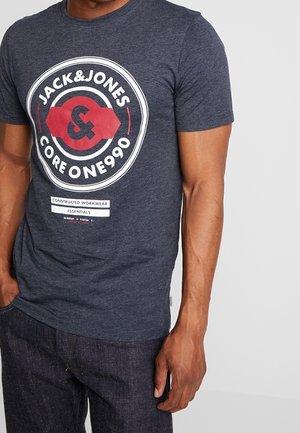 Mężczyzna noszący ciemnoniebieską koszulkę Jack & Jones z białym i czerwonym okrągłym logo oraz ciemne dżinsy, stoi z jedną ręką w kieszeni.