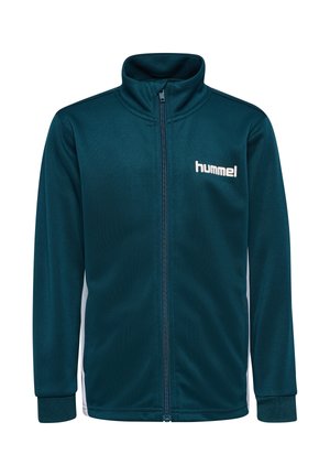 Dreng står med hænderne bag ryggen iført teal lynlåstrøje og shorts med hvide og sorte chevronstriber og hvide sneakers.