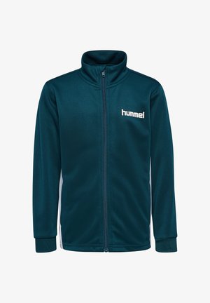 Dreng står med hænderne bag ryggen iført teal lynlåstrøje og shorts med hvide og sorte chevronstriber og hvide sneakers.