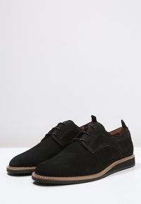 Un paio di scarpe eleganti in suede nero con lacci, suola cucita in marrone chiaro e linguette sul tallone, esposte su una superficie bianca.