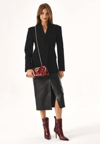Blazer nero sopra una gonna di pelle aderente con uno spacco frontale. Gli accessori includono una borsa rossa lucida e stivali alto in burgundy.