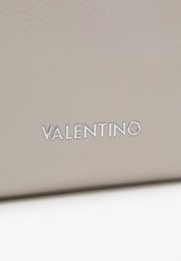 Bežs ādas soma ar teksturētu apdari, kuras apakšā ir sudraba krāsā izspiestā vārds "VALENTINO".