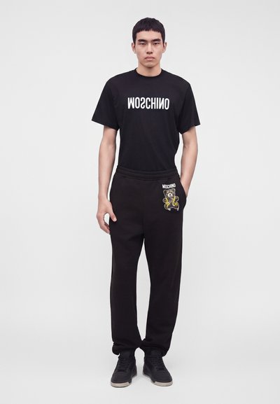 Άνδρας που φοράει μαύρο μπλουζάκι Moschino με αντίστροφο λογότυπο και μαύρες φόρμες με τσέπη αρκουδάκι, στέκεται μπροστά σε απλό λευκό φόντο.