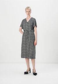 Vestido floral preto e branco com decote em V, mangas curtas, cintura ajustada e comprimento até ao joelho; combinado com sapatos de salto alto pretos.